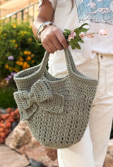 Close up of a woman in white t-shirt and jeans holding a small tote toward the camera, with a small bouquet in the same hand. The tote has an open body, solid top edge, and a decorative bow.  thumbnail