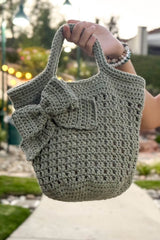 Hand holding small tote bag into frame. The bag is a pale sage green and has a decorative bow at a jaunty angle.  thumbnail