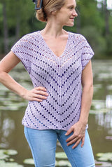 Woman in a pale purple crochet top and jeans stands on a dock in front of a pond, with lily pads.  thumbnail