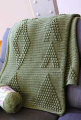 Crochet blanket in olive green draped over a grey couch. The blanket has bobbles making triangles, diamonds, and arrows on a flat background.  thumbnail