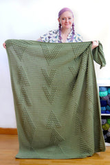 Woman holds crochet throw blanket toward camera. The blanket covers her from floor to upper chest. It is green and has raised textured designs. thumbnail