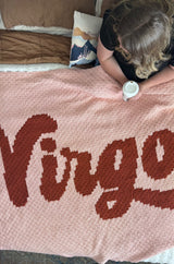 Woman sitting on a bed under a crochet afghan. The blanket is pink with deep red-orange lettering spelling the word "Virgo" thumbnail