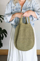 Cropped close up of crochet tote bag with short handles. The woman holding the bag is wearing a light denim jacket and white dress. The bag is crocheted with a light olive green yarn in a semi-open stitch and has short handles that are lightly stuffed.  thumbnail