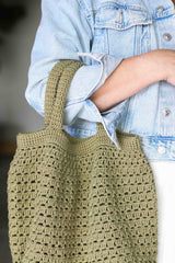 Close up of crochet bag worn in the crook of woman's elbow. The handles are lightly stuffed and the top of the bag has a solid stitch, with a more open body. It's made in a yellow olive green yarn.  thumbnail