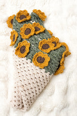 Crochet blanket wrapped up to look like a bouquet of sunflowers. The body of the blanket is cream, with a sage green border and brown and golden flowers.  thumbnail