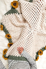 Top down view of crochet blanket open on a lap. You can see the blanket's hexagonal body done in cream, with a sage green border all around, and sunflowers on some edges.  thumbnail