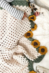 Close view of half a blanket, with a cream colored granny stitch center, sage green border, and flower appliques. The flowers have a dark brown center and golden yellow petals.  thumbnail