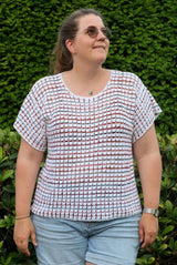 Woman standing outdoors in front of a green hedge. She wears a ponytail, sunglasses, jean shorts, and a crochet top. The top is mainly white with small colorful stripes.  thumbnail
