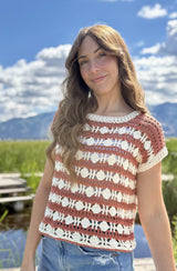 Three-quarter side view of woman wearing crochet top and denim bottoms, in front pf a pond with tall green reeds in background.  thumbnail
