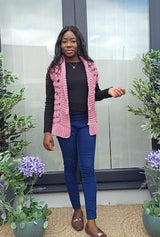 Woman stands on patio in front of glass double doors, with potted plants on either side. She wears jeans and a black top, with a pink crochet vest worn open over top.  thumbnail