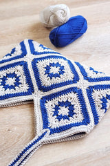 Front view of bag laying on wooden table. The top opening has a zig zag edge due to bag construction of multiple squares. Two balls of the same blue and off-white yarn are in the background.  thumbnail