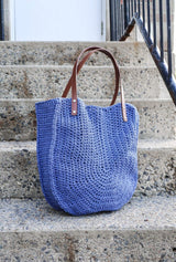 Crochet tote bag sits on cement stairs. It has dark brown leather straps and is made from a denim blue yarn. The front of the bag has a horseshoe stitch shape.  thumbnail