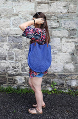 Woman in a bright coverup and shorts stands sideways to camera. She has a crochet tote bag over her shoulder, it comes from her armpit to low hip. The bag is a bright denim blue.  thumbnail