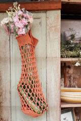 A crocheted market bag hangs on a weathered door, holding a bouquet of flowers at the top and vegetables at the bottom.  thumbnail