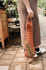Hip down view of woman in a tan dress and white sneakers holding a narrow crochet market bag, with green vegetables inside.  thumbnail