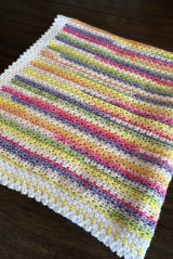 Close up of blanket including bottom order. The border is a mix of white and self-striping yarn. The body is self-striping in white, purple, pink, orange, yellow, and green.  thumbnail