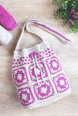 Pink and white crochet bag laid on a pale wood surface, with a spray of greenery in the upper right corner and two balls of yarn in the left corner.  thumbnail