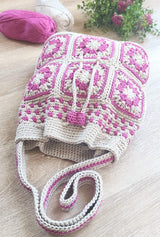 Front view of crochet bag laid on table. The bag is made of pink and cream granny squares, with a solid cream band at the top, a drawstring closure, and a strap.  thumbnail