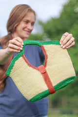 Woman holds crochet bag toward camera. The bag has a yellow body, green bottom and top handles, and a red-orange strap around the middle.  thumbnail