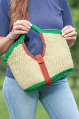 Woman holds crochet purse at jaunty angle. The purse has bright green trim at top and bottom, red-orange trim around the inner handle and as a vertical strap around the middle. The body of the bag is a straw yellow color.  thumbnail