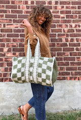 Side view of woman holding crochet duffel bag in the crook of her arm. The bag has an olive and cream checkered pattern on the body, solid olive ends, and two straps that go all the way around the middle of the bag.  thumbnail