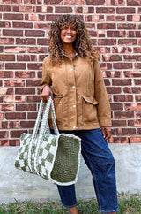 Front view of woman in brown coat and jeans holding a crochet bag. he bag has a solid green end and a checkered body.  thumbnail