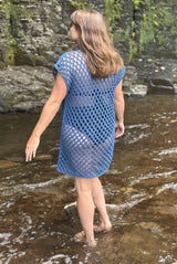 Back view of a woman standing in a stream, with a cliff face in the background. She wears a blue mesh crochet cover up over a two piece swimsuit.  thumbnail