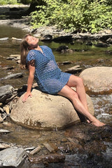 Woman lounges on a sun-kissed rock in the middle of a stream, wearing a blue mesh coverup over a swimsuit.  thumbnail