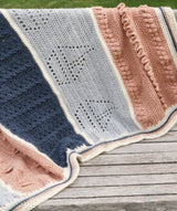 Cropped view of crochet afghan laid over wooden bench. Light blue section has eyelets in the shape of a sailboat, pale orange section has bobbles and ruffles.  thumbnail