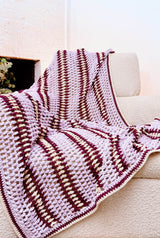 Crochet afghan draped over a cream armchair in a neutral living room. The afghan is made in pale purple, dark red-purple, and cream.   thumbnail