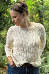 Woman standing in a forest wearing a cream knit sweater and dark blue jeans. The sweater has a geometric cabled design on the front, a ribbed collar, and plain sleeves.  thumbnail