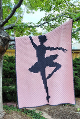 Crochet afghan draped over a tree branch. The afghan is pale pink with a ballerina figure in dark grey, with a dark grey border.  thumbnail