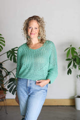 Woman stands in front of a white wall with two potted plants behind her. She wears blue jeans and a bright green turquoise crocheted pullover.  thumbnail