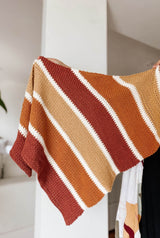 Person offscreen holds a crochet shawl in a white room. The shawl is striped in brick red, saffron orange, pale peach, and dark wine red. Each stripe is separated by a smaller stripe of cream.  thumbnail