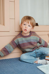 Young boy sits on a deck in jeans and a striped sweater. The sweater is striped in blocks of brown, orange, blue, and pale green.  thumbnail