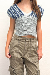 Cropped straight-on front view of crochet top worn with khaki pants. The top has a v-neck, solid blue body, and wide shoulders with vertical textured stripes in blue and darker blue.  thumbnail