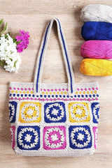 Crochet tote bag laid on wooden table top, with four balls of yarn in the upper right corner and flowers on the left side of the background.  thumbnail