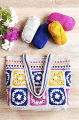 Top down view of crochet tote bag on wooden table top. The bag handles are folded down, and there are flowers and yarn skeins above the bag on the table top.  thumbnail