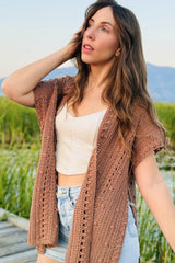 Woman in a tank top and shorts wears an open cardigan crocheted in a tan yarn. She is standing on a boardwalk in a wetland area, with a mountain in the background.  thumbnail
