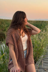 Front side view of woman sitting on the edge of a boardwalk in a wetland area, looking over her left shoulder. She wears an open short sleeve cardigan over a tank top and jean shorts.  thumbnail