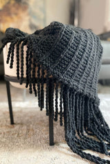 Knit throw in charcoal grey draped over a white armchair in a modern living room. The throw has a textured pattern and twisted fringe.  thumbnail