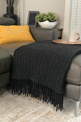Knit throw in charcoal grey folded neatly over a paler grey leather couch. There is a yellow throw pillow, side table with decorative objects in the background.  A circular tray with a coffee mug sits on top of the afghan.  thumbnail