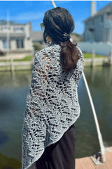 Side back view of woman on a dock wearing a knit shawl with an intricate lace pattern. The shawl is knit in a bulky weight pale blue-grey yarn.  thumbnail