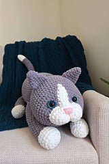 Large crocheted cat sits in a cream armchair with a dark blue afghan in the background. The cat is grey with a white nose, paws, and tail tip. It has a pink nose and blue plastic eyes.  thumbnail