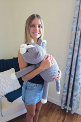 High angle view of woman holding stuffed crochet cat. The head rests on her shoulder and the body goes to her waist. The legs and tail are splayed to the sides.  thumbnail