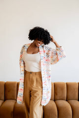 Woman wearing a white tank top and beige pants stands in front of a brown velvet sofa. She also wears a long crochet cardigan in a granny stitch pattern with a ribbed front band.  thumbnail