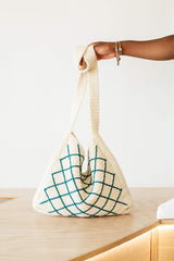 A hand from a person offscreen holds the long strap of a crochet tote bag, which rests on a wooden countertop.  thumbnail