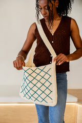 Woman in a vest top and jeans wears a crochet tote bag. The bag has a long strap and a wide front panel with narrow sides. It is made in a cream yarn with a diamond pattern in a dark jade worked over top.  thumbnail