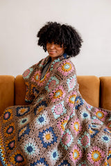 Woman sitting on a plush couch wrapped in a granny square blanket. The blanket is made of many squares in varying color arrangements.  thumbnail