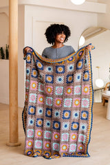 Woman in a grey top holds up a crochet afghan, covering her from chest to floor. The afghan is made of many granny squares arranged in subtle stripes basked on color combination. The blanket has a border all the way around.  thumbnail